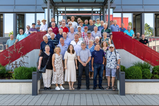Gruppenfoto von mehreren älteren Menschen auf einer Treppe vor einem modernen Gebäude des Bauunternehmens KASSECKER. Einige Personen tragen Hüte wegen des sommerlichen Wetters