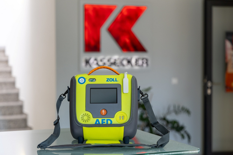 Gelber Defibrillator mit Tragegurt auf Glasfläche vor einem an der Wand hängenden KASSECKER-Logo