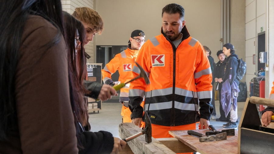 Mehrere Personen in einer Werkhalle, zwei Männer in orangefarbenen Arbeitsjacken mit reflektierenden Streifen und KASSECKER Logo, eine Person hält einen Hammer über einem Holzstück