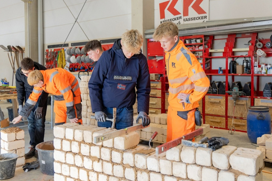 Vier Jugendliche, zwei davon in KASSECKER Arbeitskleidung, bauen in einer Halle eine Mauer und eine Säule aus Ziegelsteinen in einer Werkstatt