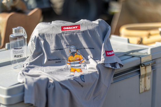 T-Shirt mit Aufdruck einer grafisch erstellten Person in Schutzkleidung und Warnschutzjacke sowie Texten zu Schutzhelm, Schutzbrille, Warnschutzjacke und Schutzweste. Daneben steht eine Trinkflasche mit Aufdruck. Beide sind mit dem Schriftzug "OBACHT" gebrandet.