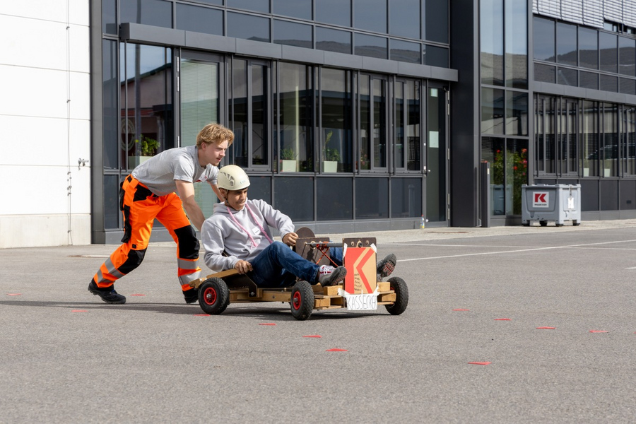 Ein Mann in grauem T-Shirt und oranger Arbeitskleidung schiebt einen anderen Mann mit Helm, grauem Pullover und Jeans in einem selbstgebauten Seifenkistenwagen auf einem Asphaltplatz. Die Seifenkiste ziert ein KASSECKER-Logo.