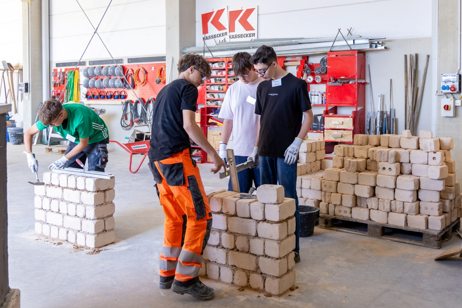 Vier junge Männer in Werkstatt mit Ziegelsteinen bauen Mauern, im Hintergrund Werkzeuge und KASSECKER-Logo an der Wand