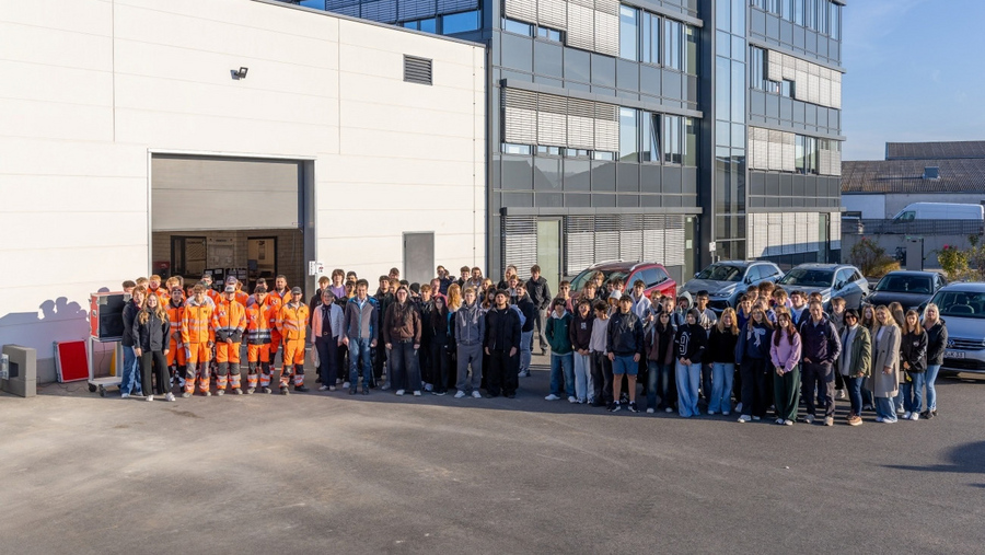Große Gruppe von Menschen, darunter mehrere Personen in orangefarbenen Arbeitsanzügen, steht vor einem Industriegebäude mit großen Fenstern und geparkten Autos
