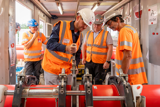 Drei Männer des Bauunternehmens Franz KASSECKER GmbH mit Schutzhelmen und orangefarbenen Warnwesten arbeiten an einer roten Rohrleitung in einem Container