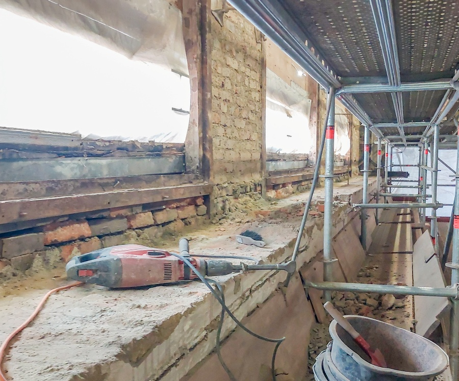 Baustelle mit Gerüst, Mauer mit freigelegtem Mauerwerk, Elektrowerkzeug auf Fensterbank und Eimer mit Schutt auf Boden