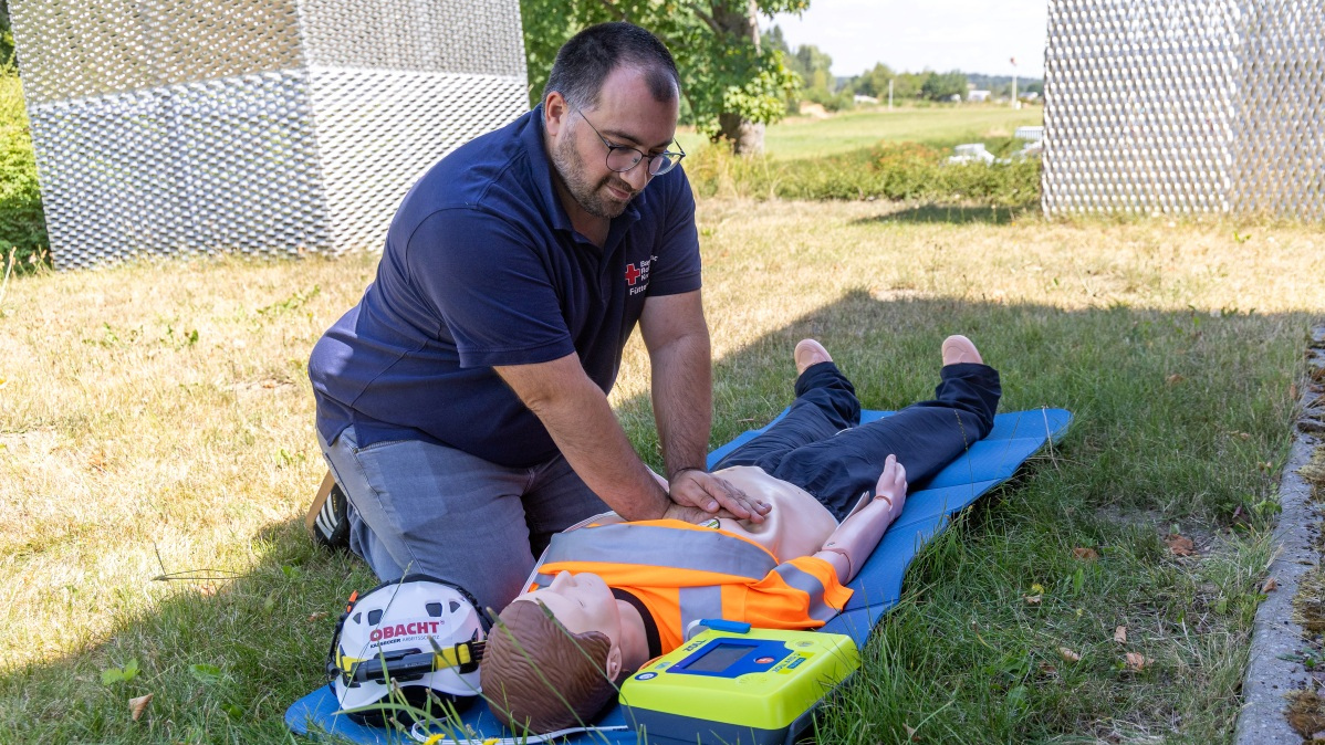 Mann in blauem Poloshirt mit Brille führt Herzmassage an Reanimationspuppe in orangefarbener Warnweste auf blauer Matte im Freien durch, daneben liegen Helm und Defibrillator.