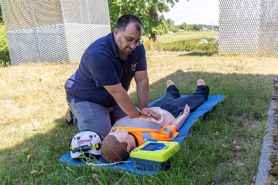 Mann in blauem Poloshirt mit Brille führt Herzmassage an Reanimationspuppe in orangefarbener Warnweste auf blauer Matte im Freien durch, daneben liegen Helm und Defibrillator.