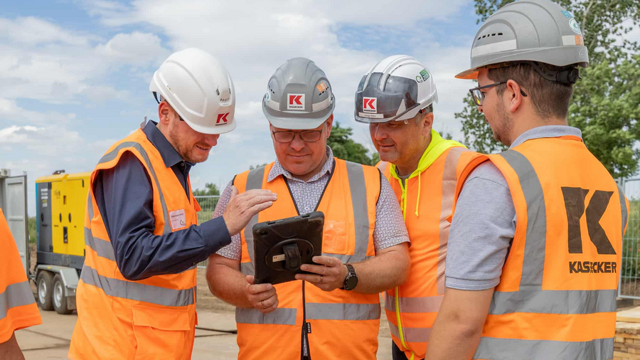 Vier Kassecker Mitarbeiter auf einer Baustellen schauen in ein Tablet.