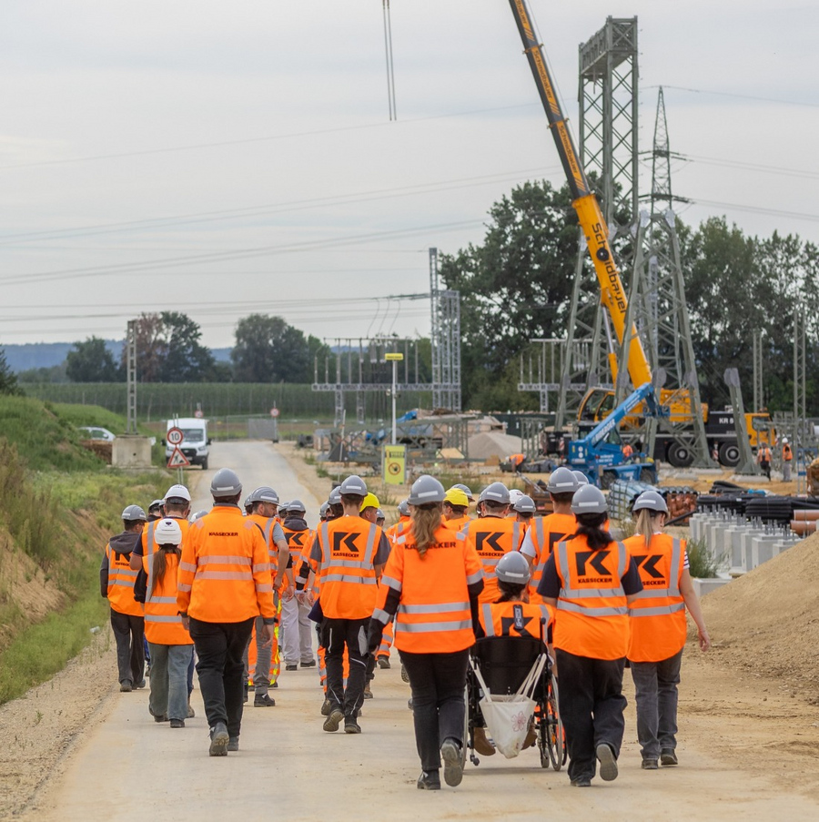 Gruppe von Menschen mit orangen Warnwesten und Schutzhelmen geht auf einer Baustelle mit Kränen und Strommasten