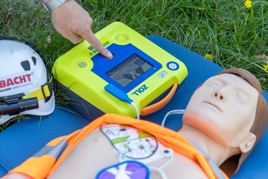 Hand drückt Knopf an gelbem Defibrillator neben liegender Reanimationspuppe auf Wiese.