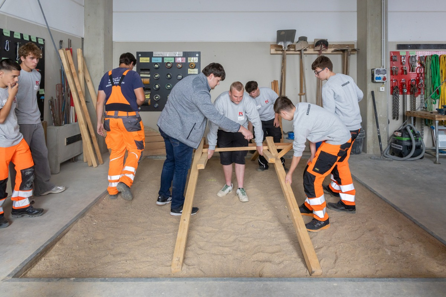 Gruppe junger Männer in Arbeitskleidung, teils mit KASSECKER-Logo, bauen ein Holzgestell in einer Werkstatt mit Werkzeugen an der Wand