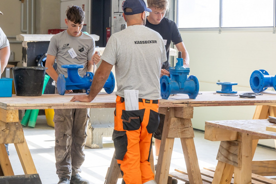 Zwei Jugendliche und ein Mann in Werkstatt mit blauen Rohrarmaturen auf Holztischen, der Mann trägt Shirt mit Aufdruck KASSECKER und orangefarbene Arbeitshose