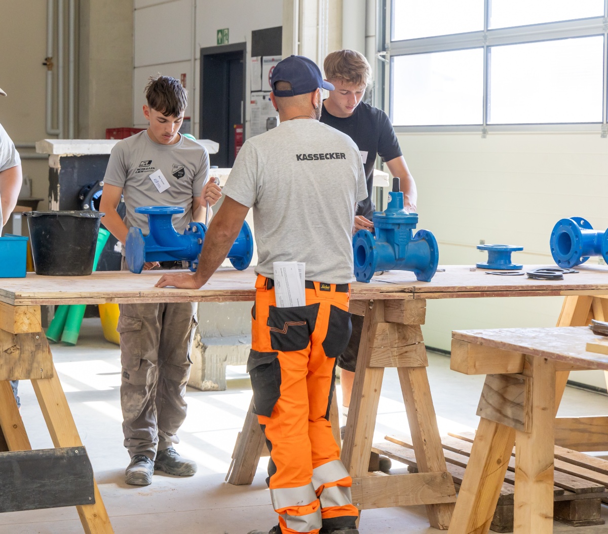 Zwei Jugendliche und ein Mann in Werkstatt mit blauen Rohrarmaturen auf Holztischen, der Mann trägt Shirt mit Aufdruck KASSECKER und orangefarbene Arbeitshose