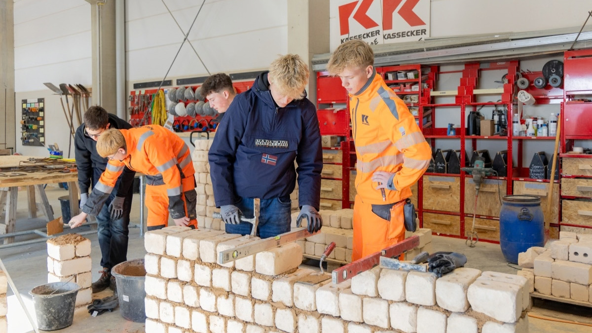 Vier Jugendliche, zwei davon in KASSECKER Arbeitskleidung, bauen in einer Halle eine Mauer und eine Säule aus Ziegelsteinen in einer Werkstatt